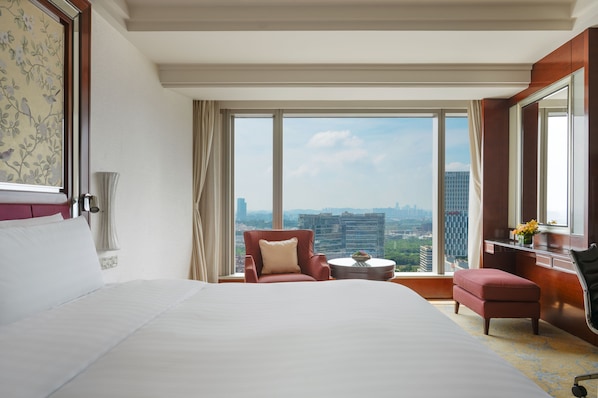 A hotel room with a large bed, a red armchair, a small table, and a view of the cityscape through large windows.