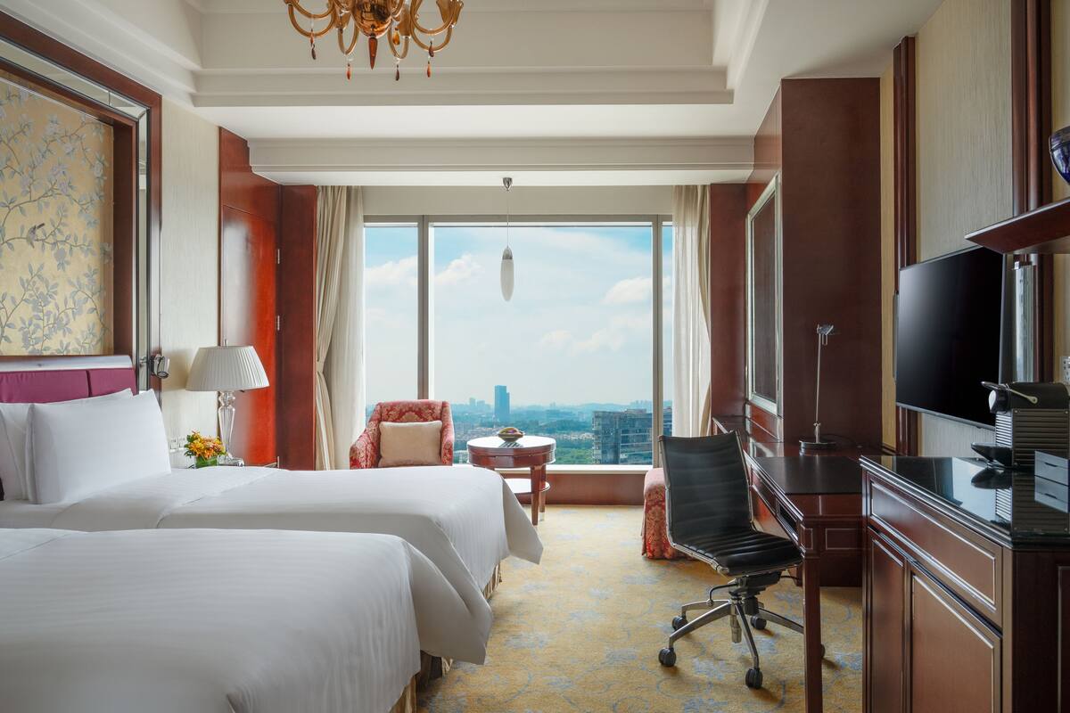 A hotel room with two beds, a desk, a TV, and a view of the city.