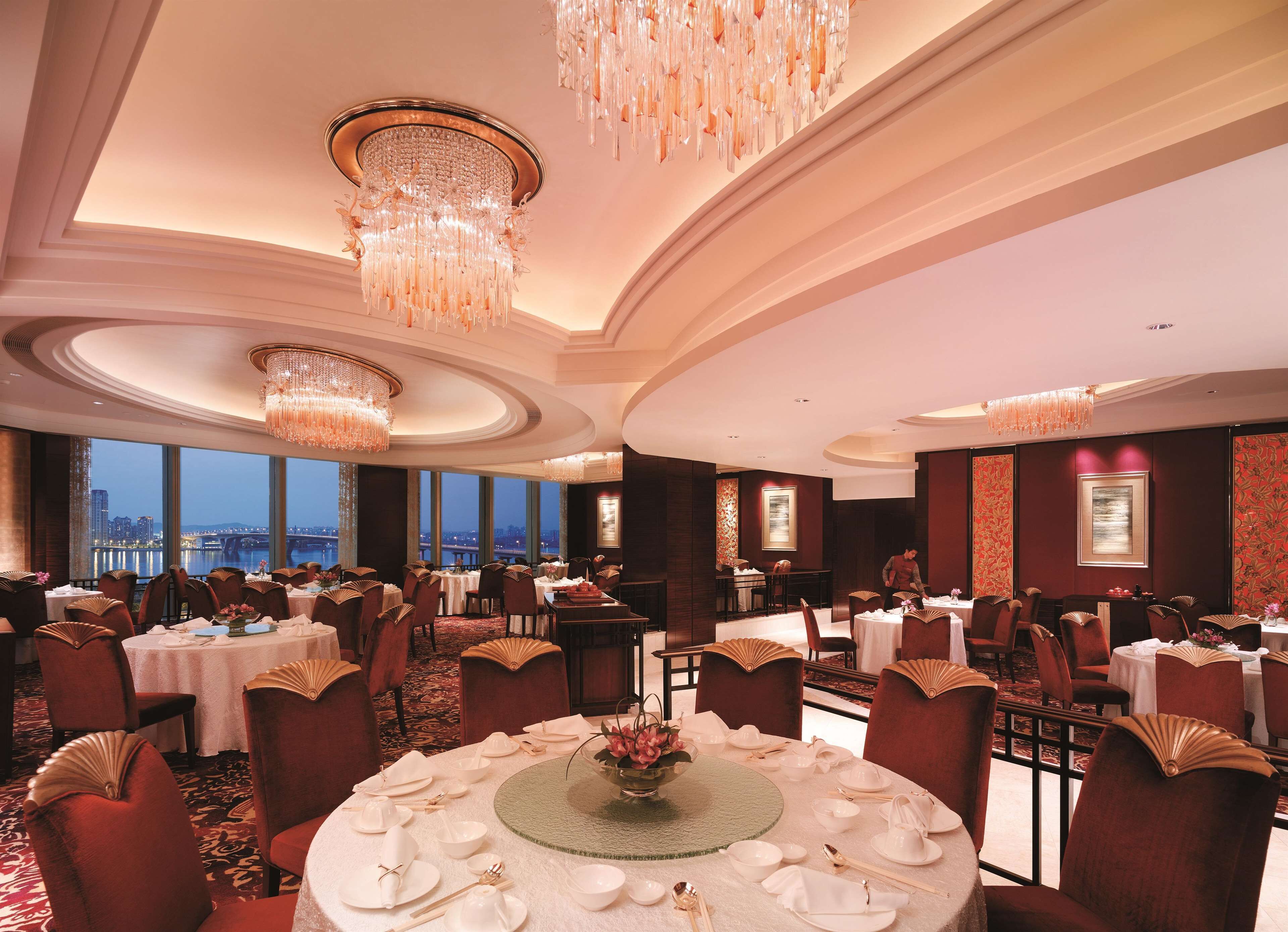 Un espace de restauration luxueux, avec de nombreuses tables dressées pour le service, d’élégants lustres et une vue sur le paysage urbain à travers de grandes fenêtres.