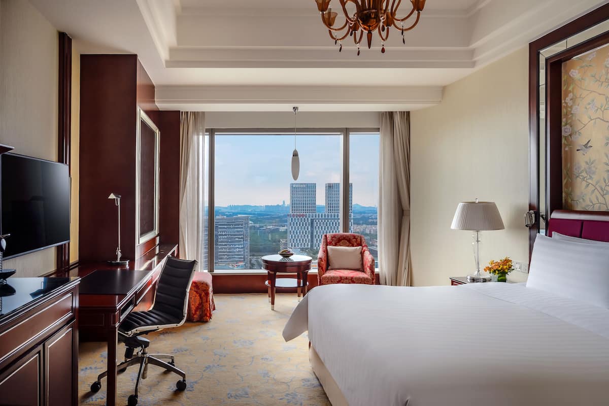 A hotel room with a large bed, a desk with a chair, a television, a small table with a lamp, and a view of the cityscape.