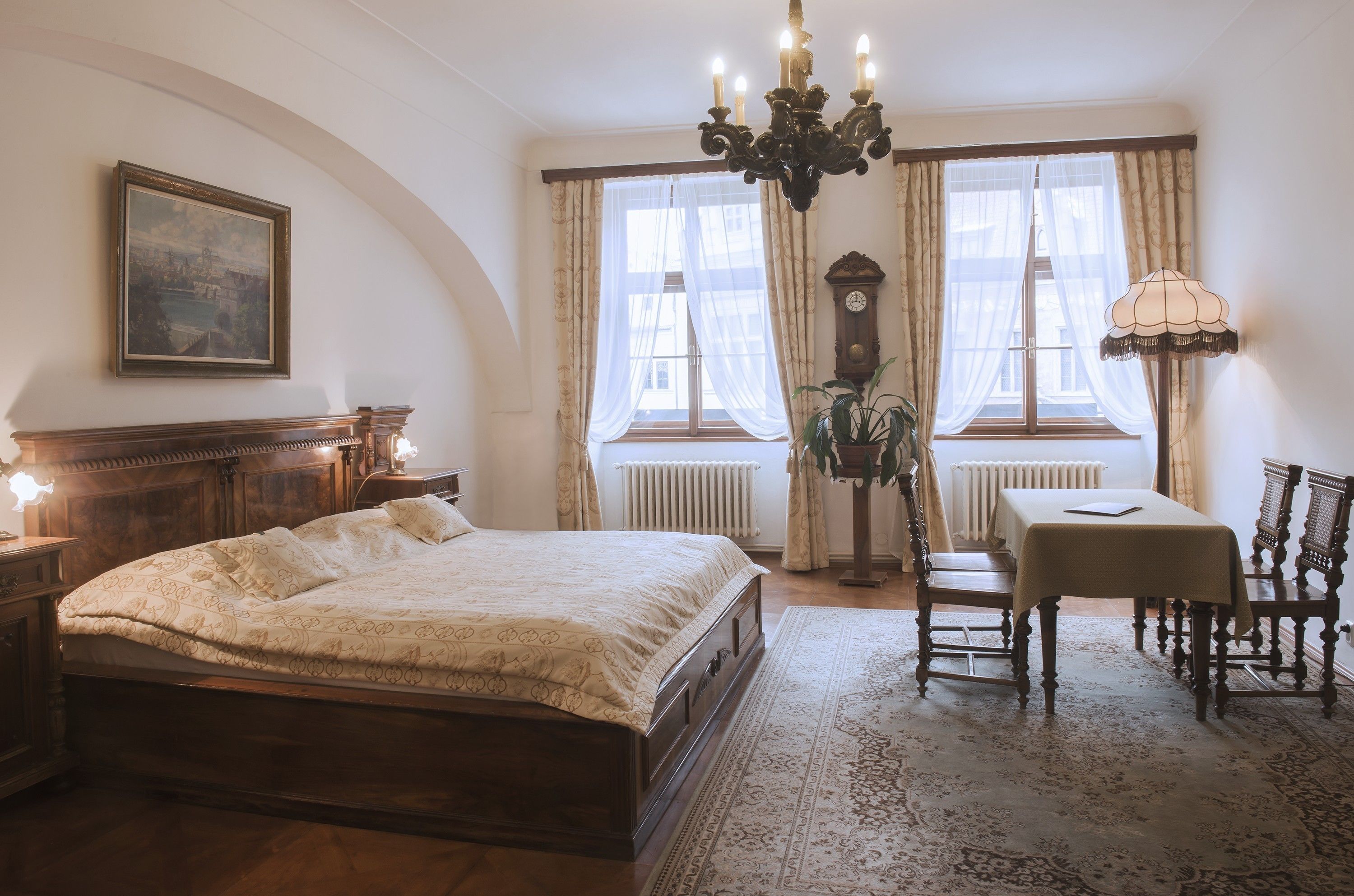 Ein Schlafzimmer mit einem großen Bett, einer Kristallleuchter, einem Tisch mit Stühlen und Blick auf die Stadt durch das Fenster.