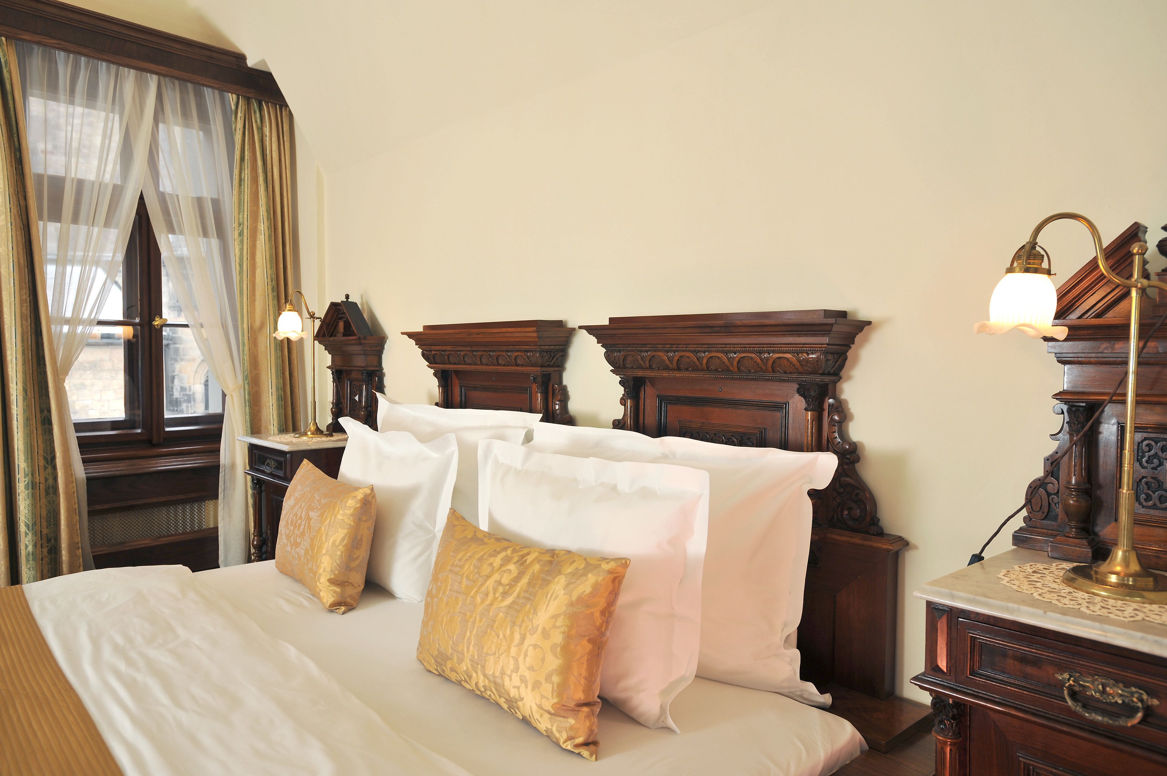 A neatly made bed with white and gold pillows, wooden headboard, and a nightstand with a lamp.
