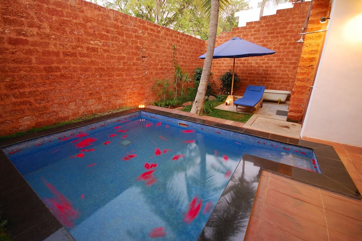 Una piscina con fuentes de agua rojas, un muro de ladrillo y un área de descanso con una sombrilla azul.