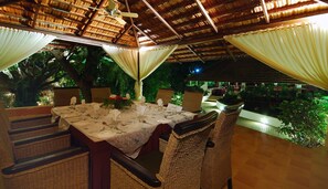 An outdoor dining area with wicker chairs and a table set for two, surrounded by lush greenery and a thatched roof structure. - The Windflower Resorts and Spa (Mysore)