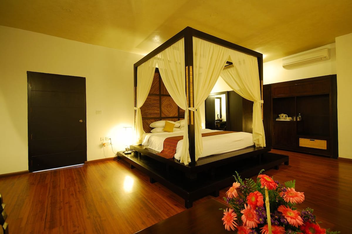 Una cama con dosel y cuatro columnas, con estructura de madera y ropa de cama blanca, un suelo de madera oscura, una mesita con un arreglo floral y un guardarropa de madera oscura.