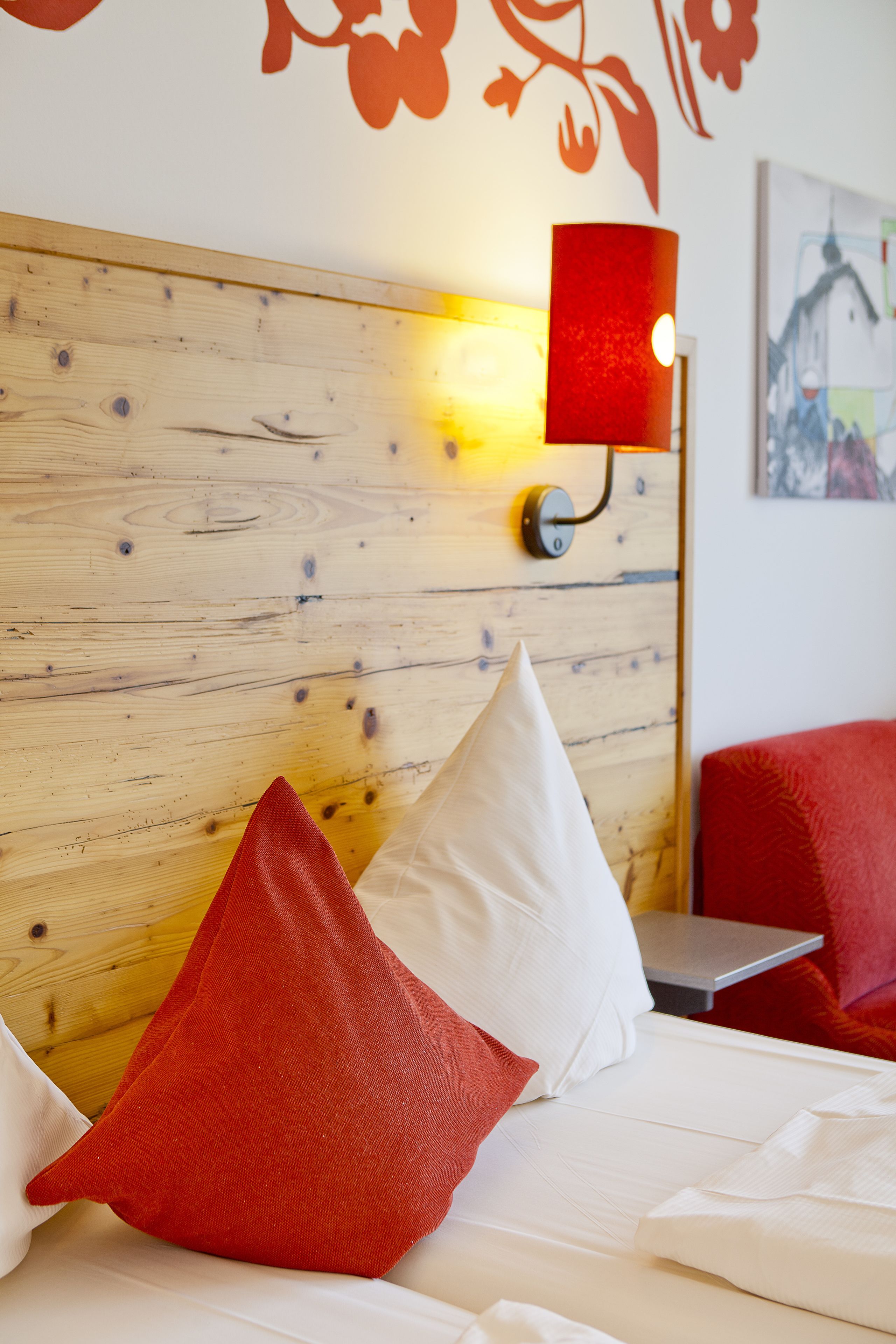 A hotel room with a wooden headboard, a red and white pillow, a red lamp, and a painting on the wall.