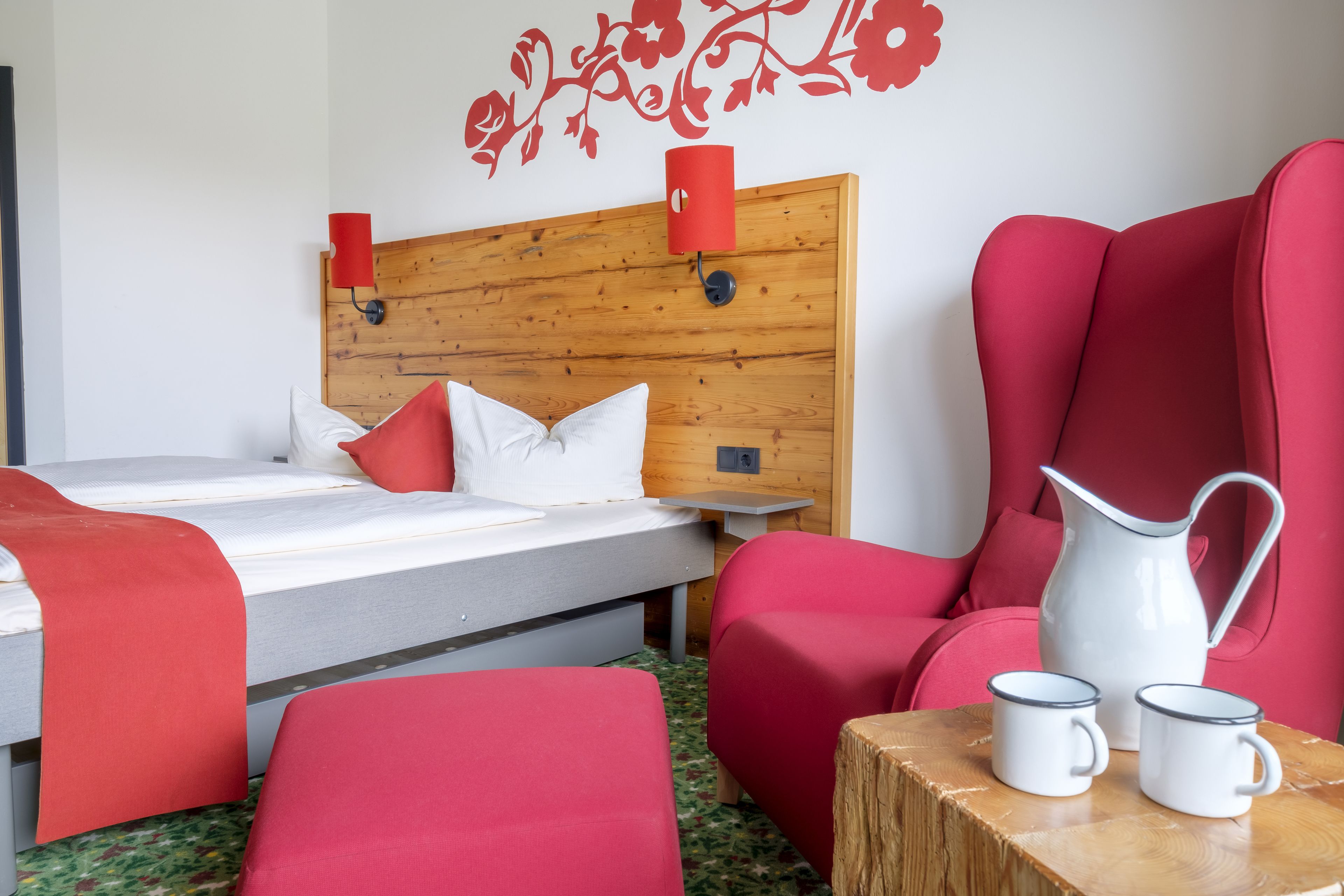A hotel room with a wooden headboard, a bed with red and white bedding, a red armchair, a wooden side table with two white mugs, and a red wall decoration.