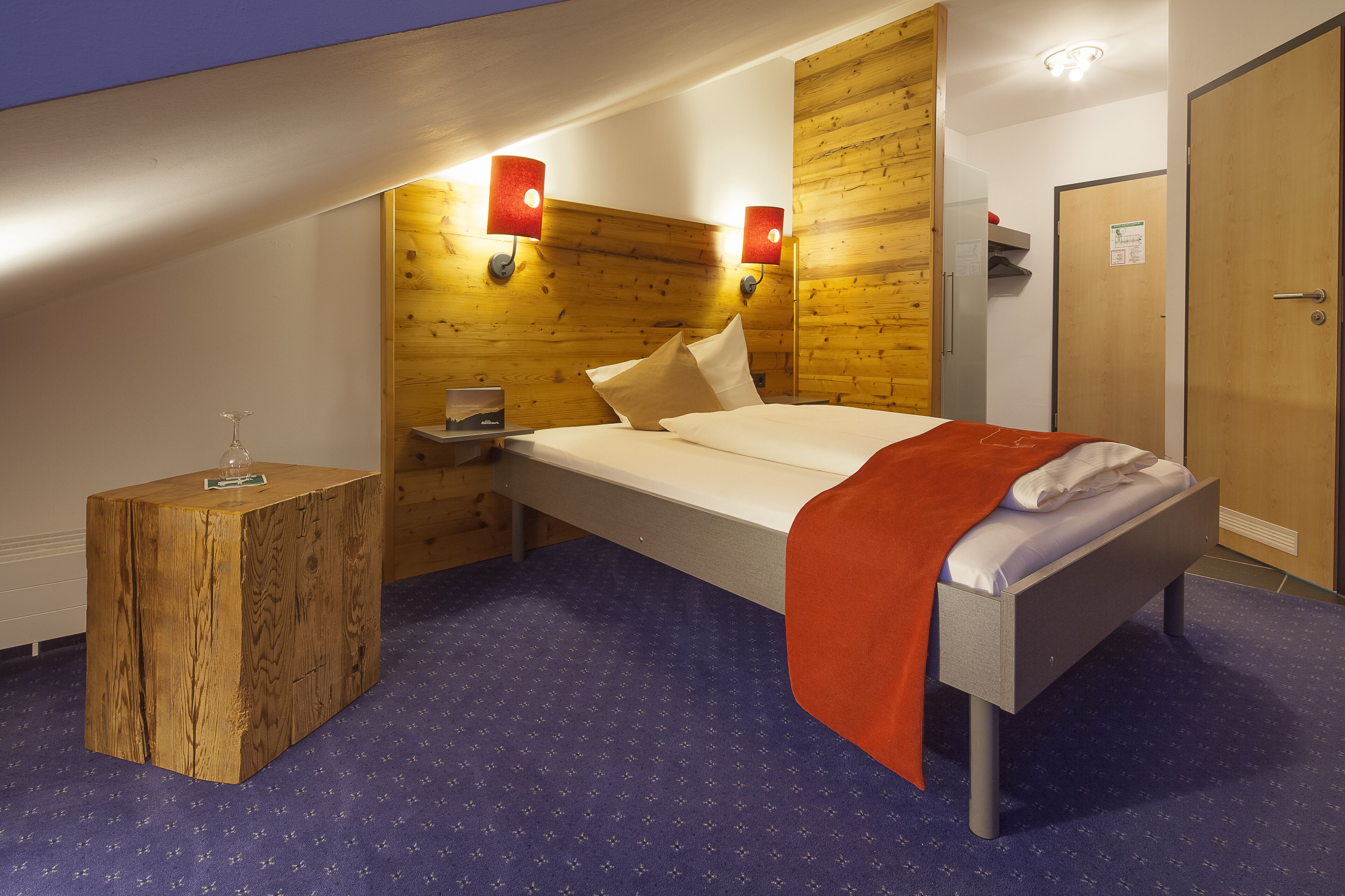 Een hotelkamer met een houten hoofdbord, een bed met een rode deken, een klein houten nachtkastje en een nachttafeltje met een lamp.