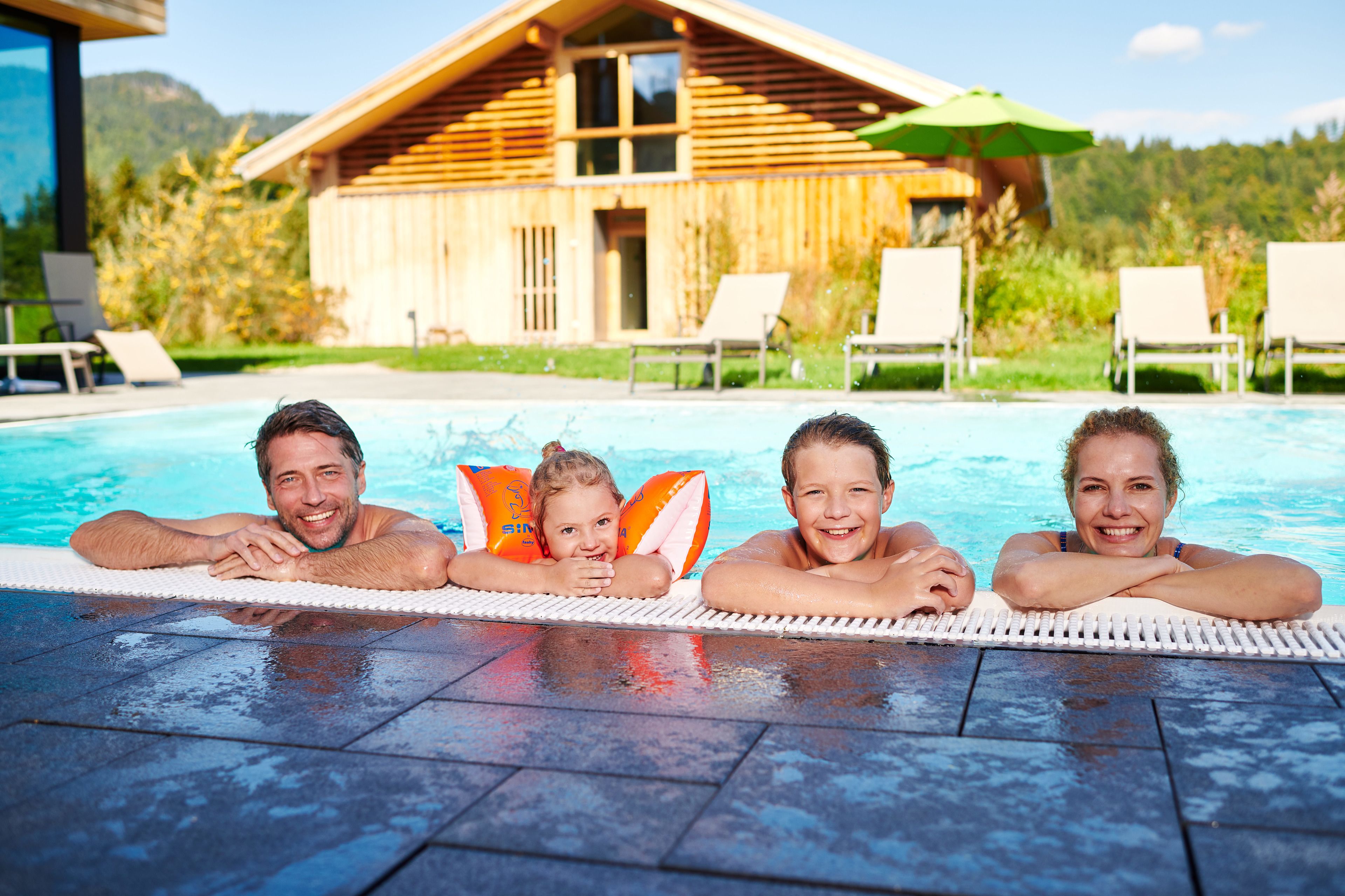 A family of four relaxing by a pool with a wooden cabin in the background.
