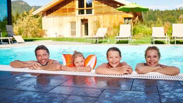 A family of four relaxing by a pool with a wooden cabin in the background.