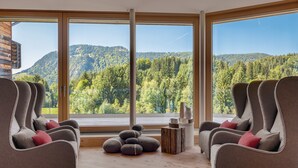 A room with large windows offering a view of a forested mountain. There are two sofas with cushions and a small wooden table with a vase and a candle. - Hotel Oberstdorf (Oberstdorf)