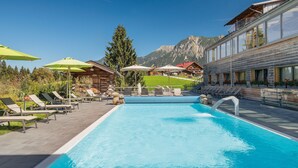 A pool area with lounge chairs and umbrellas, surrounded by wooden buildings and a mountain backdrop. - Hotel Oberstdorf (Oberstdorf)