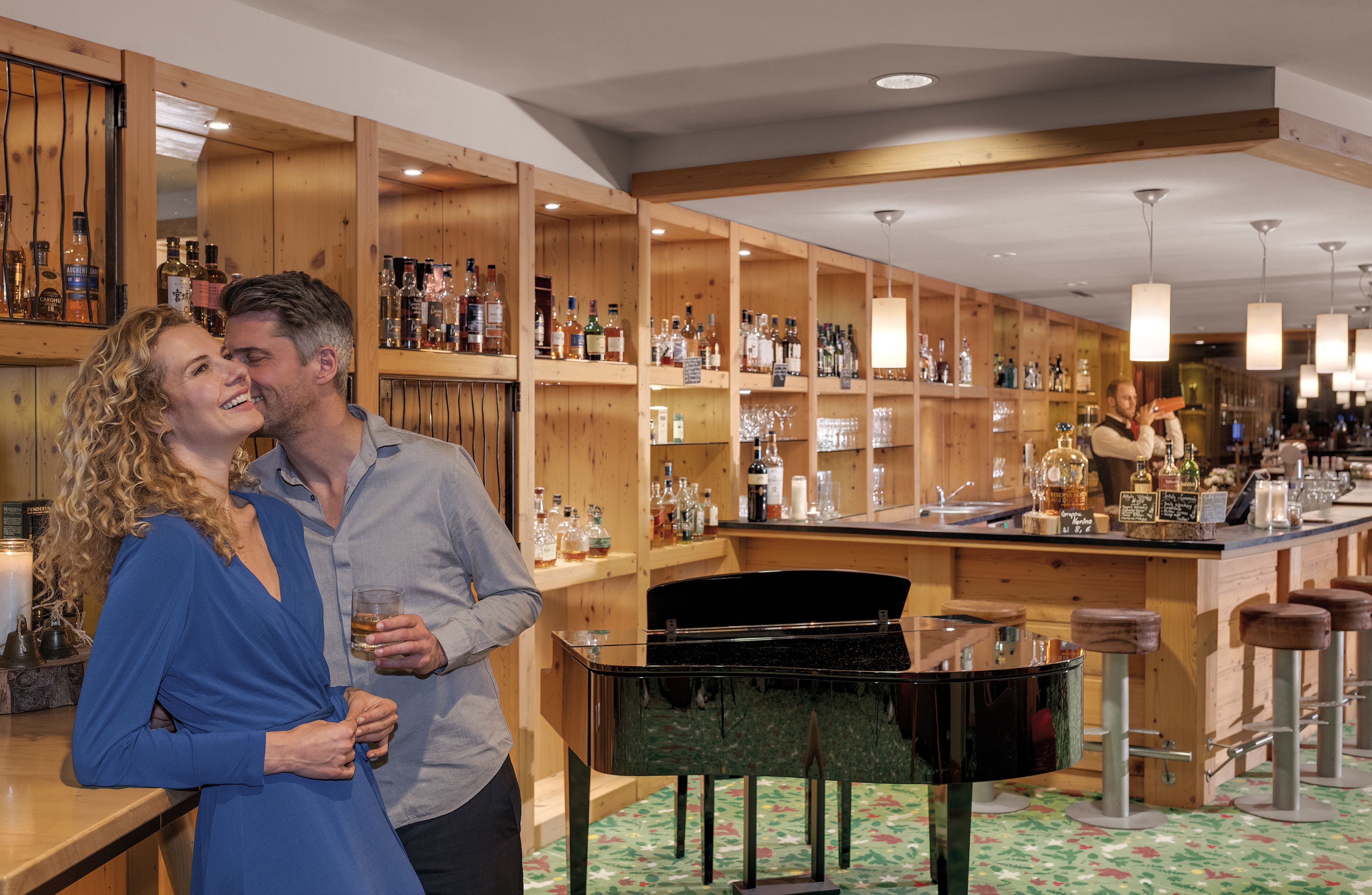 Una zona de bar con estantes de madera llenos de botellas y vasos, un piano y taburetes de bar.