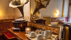 Une chambre d’hôtel avec un comptoir en bois, un gramophone, un miroir, un lit avec un oreiller et une bouilloire.