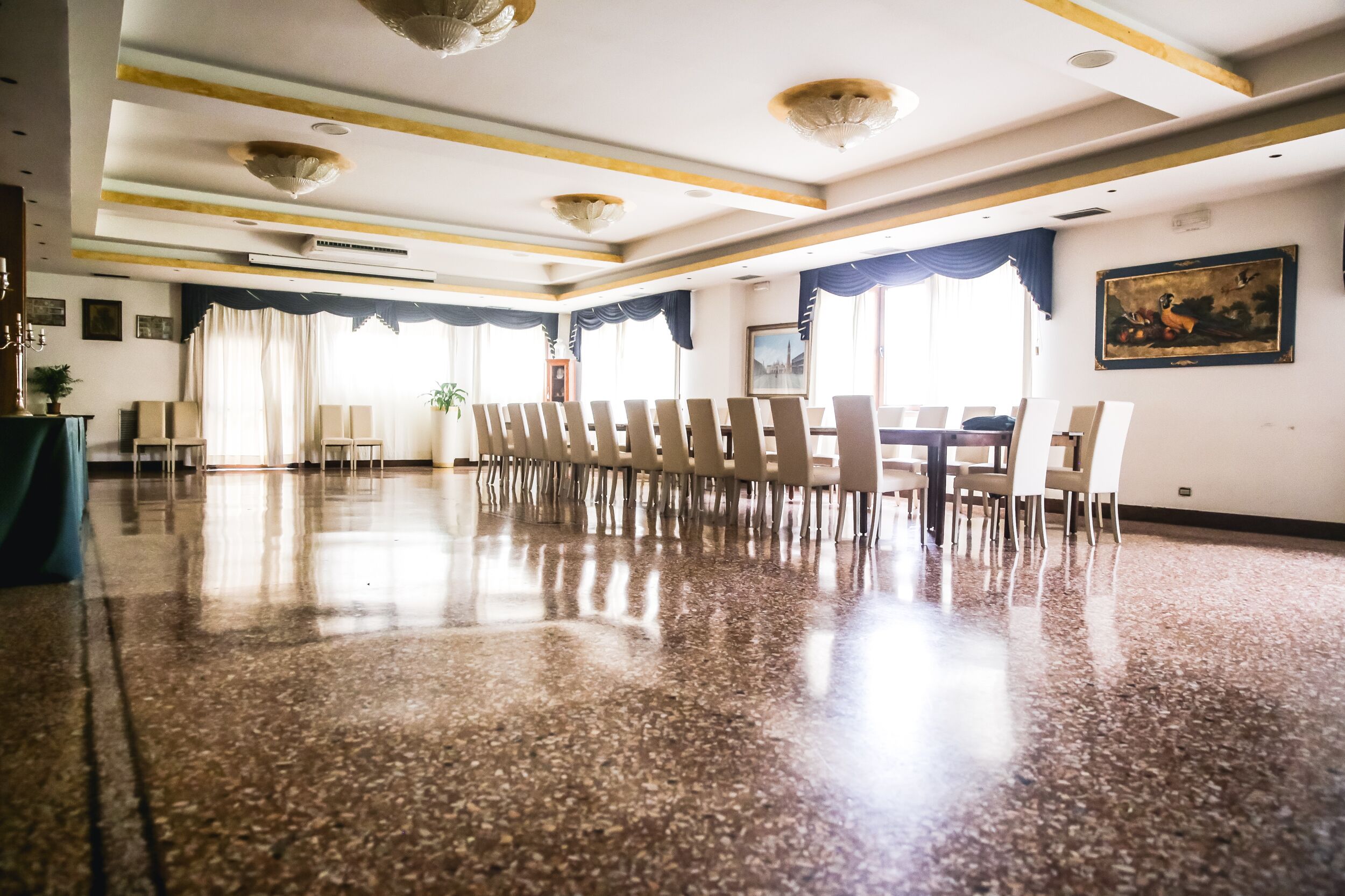 A spacious room with a polished floor, a long table with chairs, and a large window with curtains.