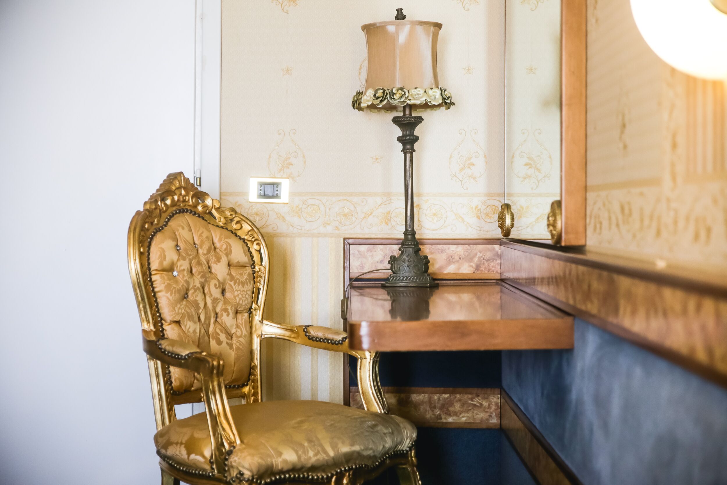 A room with a golden ornate chair, a wooden side table, and a lamp with a beige shade.