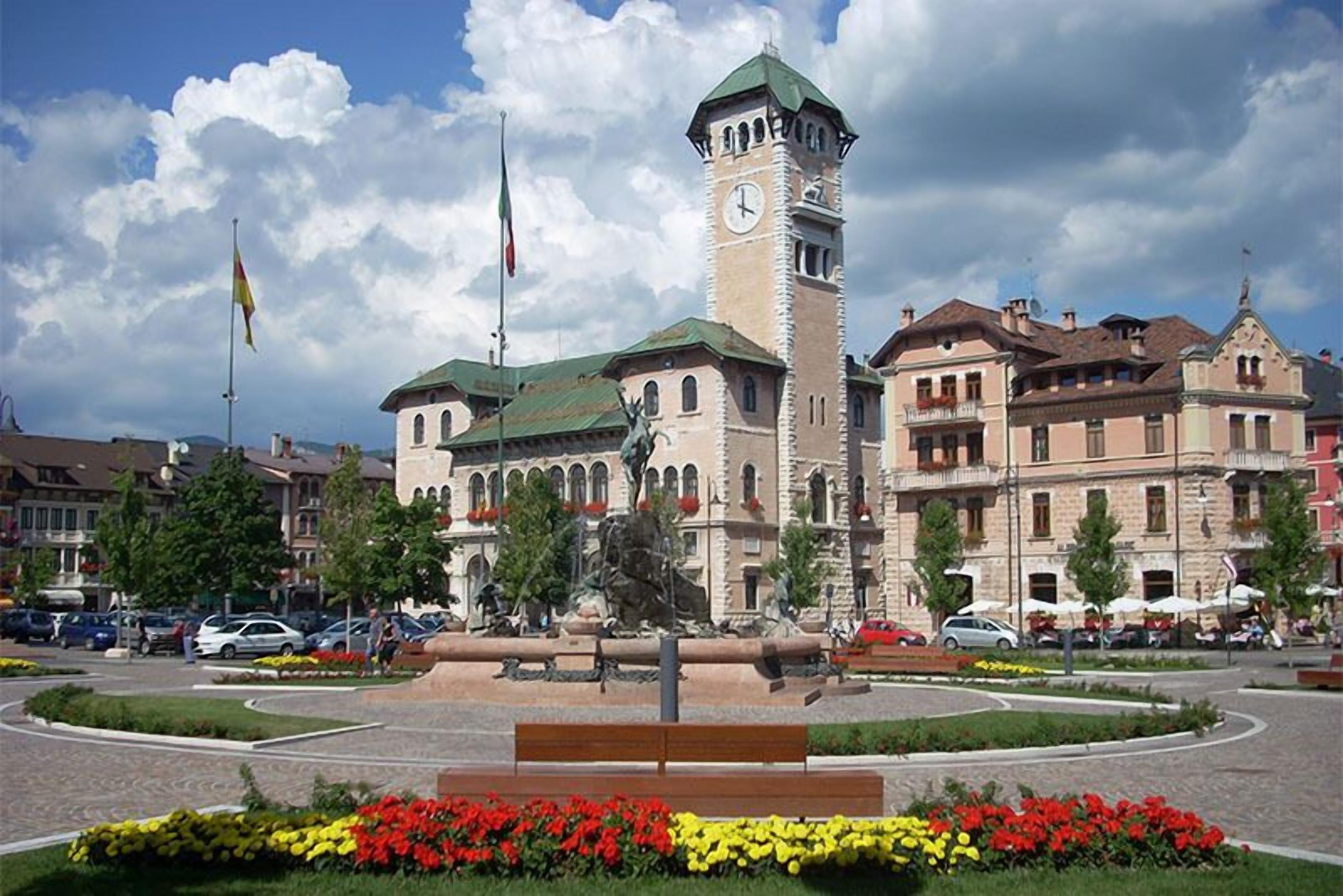 Un edificio storico con una torre dell'orologio, circondato da una piazza curata con panchine e aiuole fiorite.