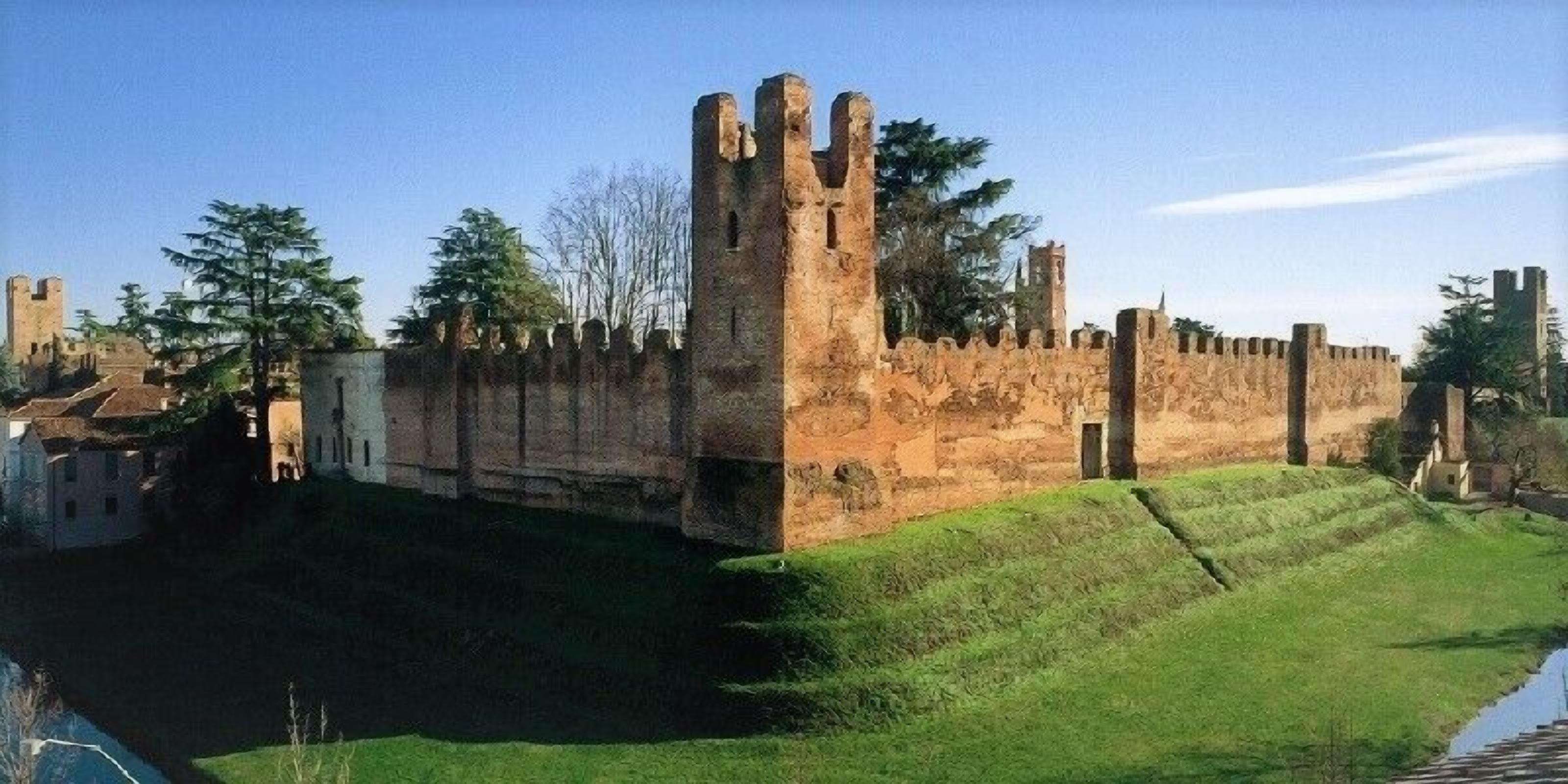 Un mur de pierre ancien, surmonté d’une haute tour, entouré d’arbres et bordé d’une zone herbeuse.