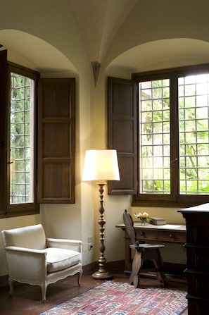 A room with a wooden desk, a chair, a lamp, and a window with a view of greenery.