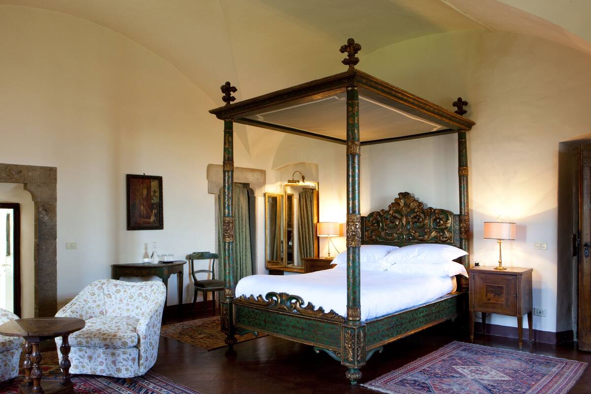 A four-poster bed with a green frame and white bedding, a wooden nightstand, a chair with floral upholstery, and a patterned rug on the floor.