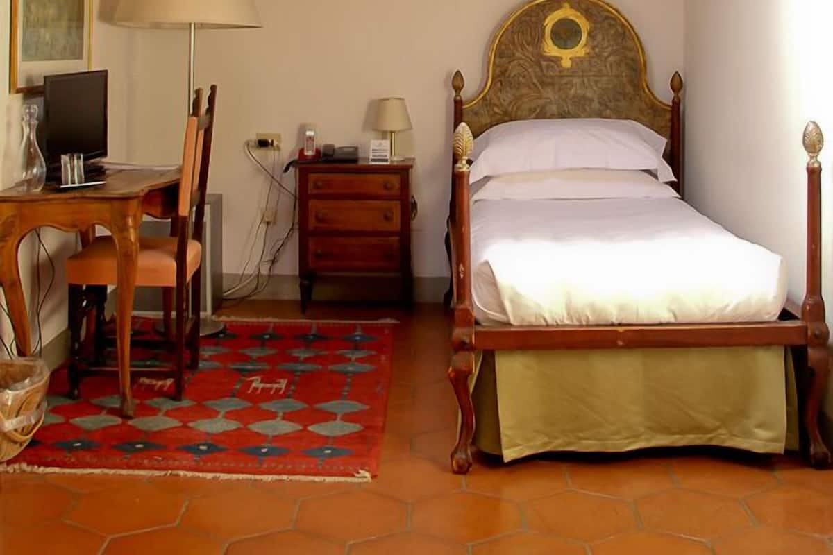 A bedroom with a wooden ceiling, a bed with a decorative headboard, a desk with a chair, a lamp, and a red patterned rug.