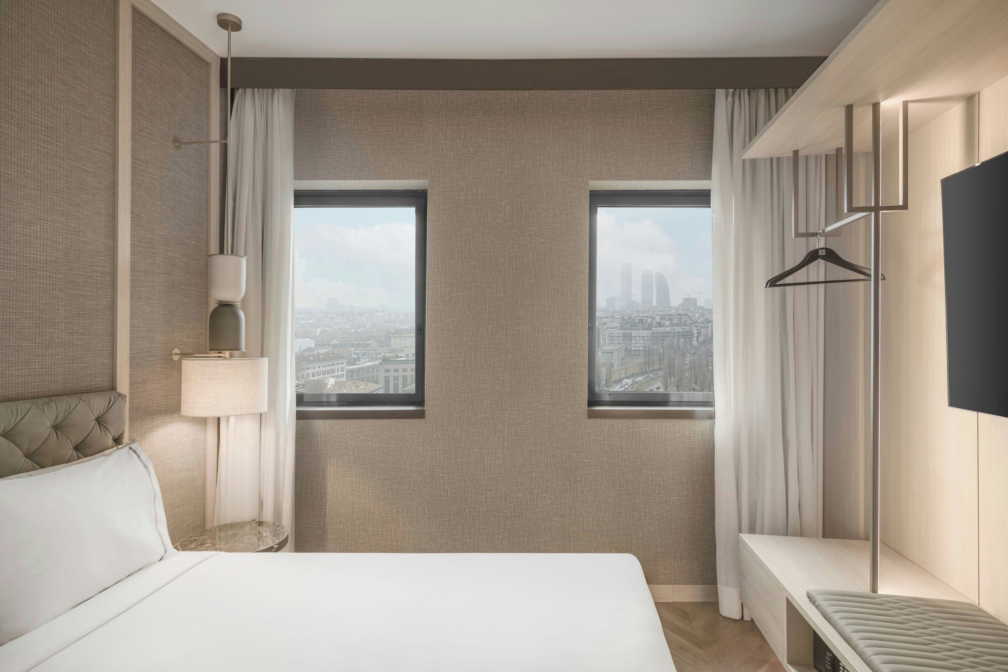 A hotel room with a bed, a bedside table, a lamp, a TV, and two windows offering a city view.