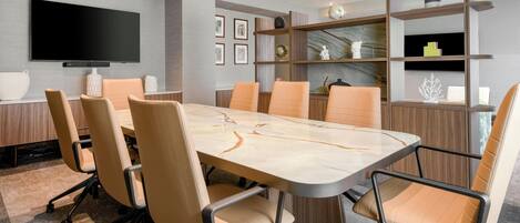 Una sala de conferencias moderna con una gran mesa, seis sillas, un televisor de pantalla plana y estanterías empotradas con objetos decorativos.