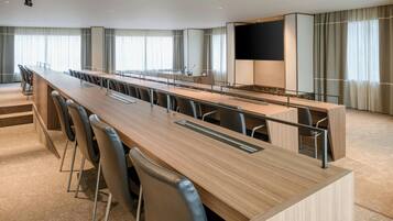 A modern conference room with a long table, chairs, a projector screen, and a large window with curtains.