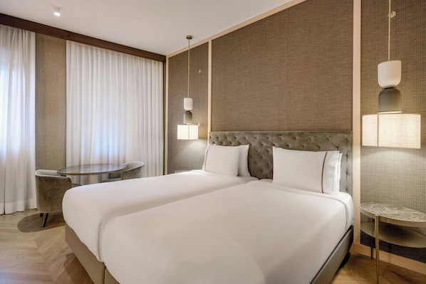 Quarto de hotel com uma cama grande, uma mesa redonda e uma luminária fixada na parede.