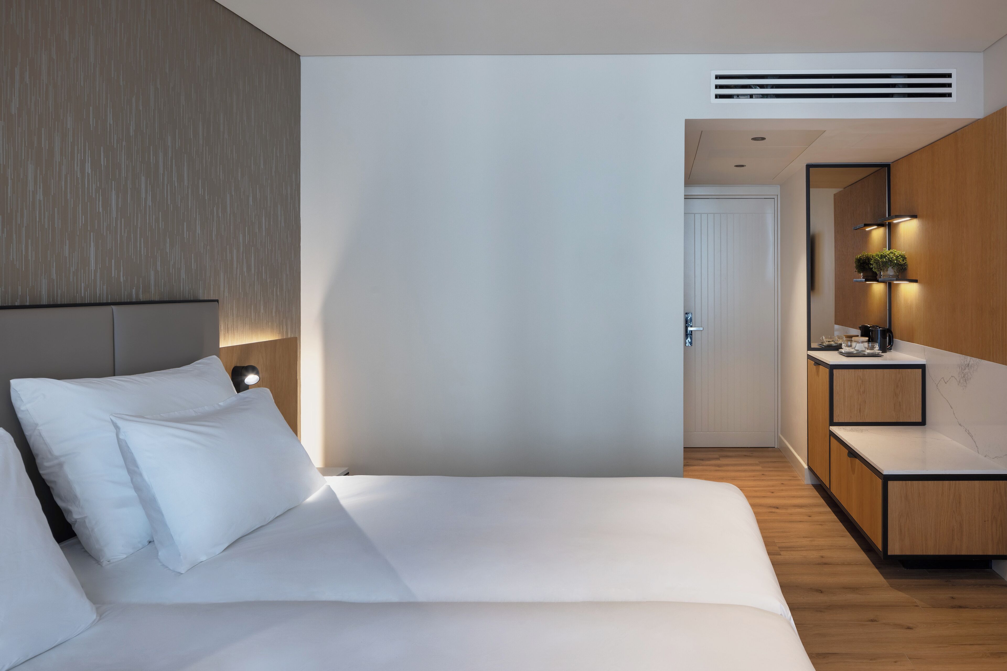 A modern hotel room with a large bed, a wooden headboard, a bedside table with a lamp, and a small bathroom area with a mirror and sink.