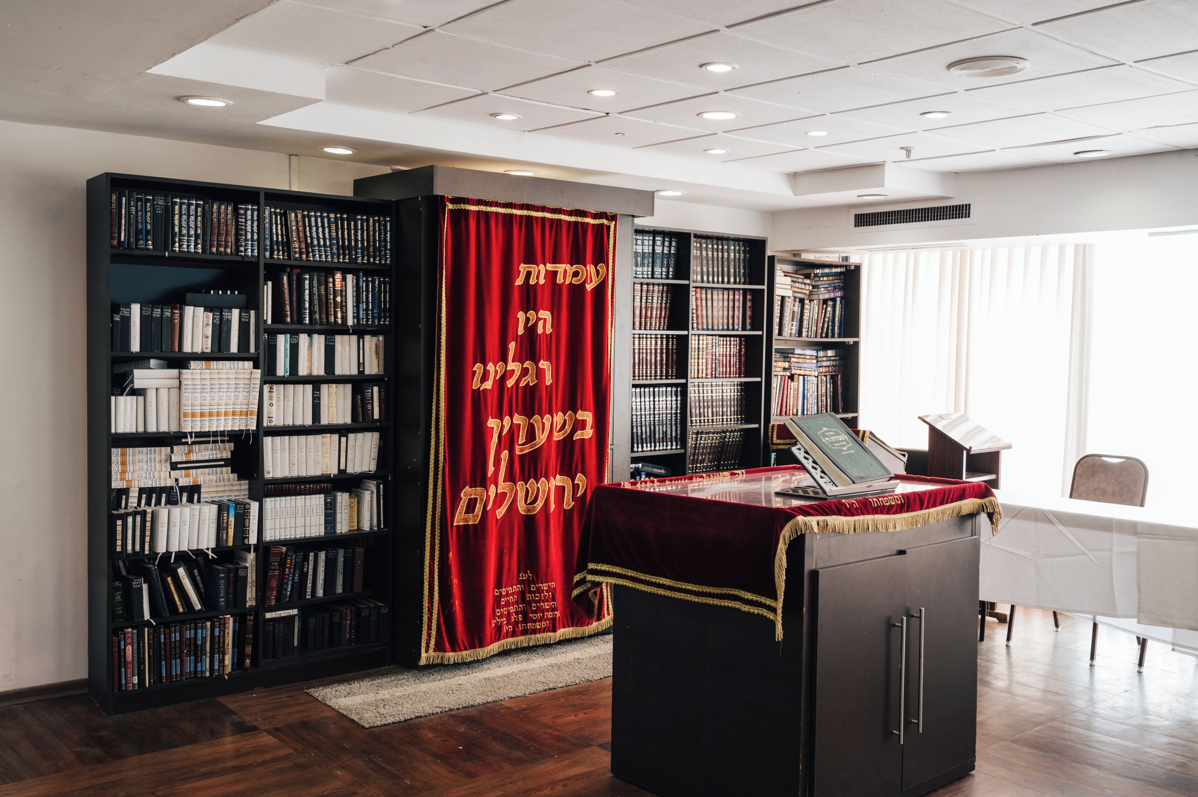 Una biblioteca con estanterías, una cortina roja con texto hebreo y un ordenador portátil sobre un escritorio.