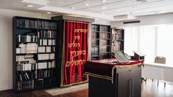 Una biblioteca con estanterías, una cortina roja con texto hebreo y un ordenador portátil sobre un escritorio.