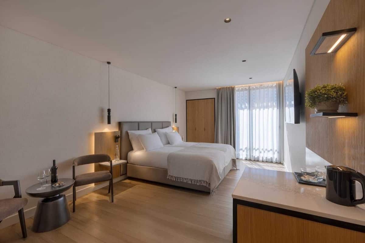 Une chambre d’hôtel moderne dotée d’un grand lit, de tables de chevet, d’un coin salon avec un fauteuil, d’une petite table ronde, d’un téléviseur fixé au mur et d’une grande fenêtre avec des rideaux.