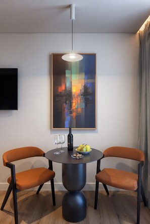Un espace repas moderne avec une table ronde, deux chaises orange, une bouteille noire et un saladier de fruits. Une peinture abstraite aux couleurs vives est accrochée au-dessus de la table.