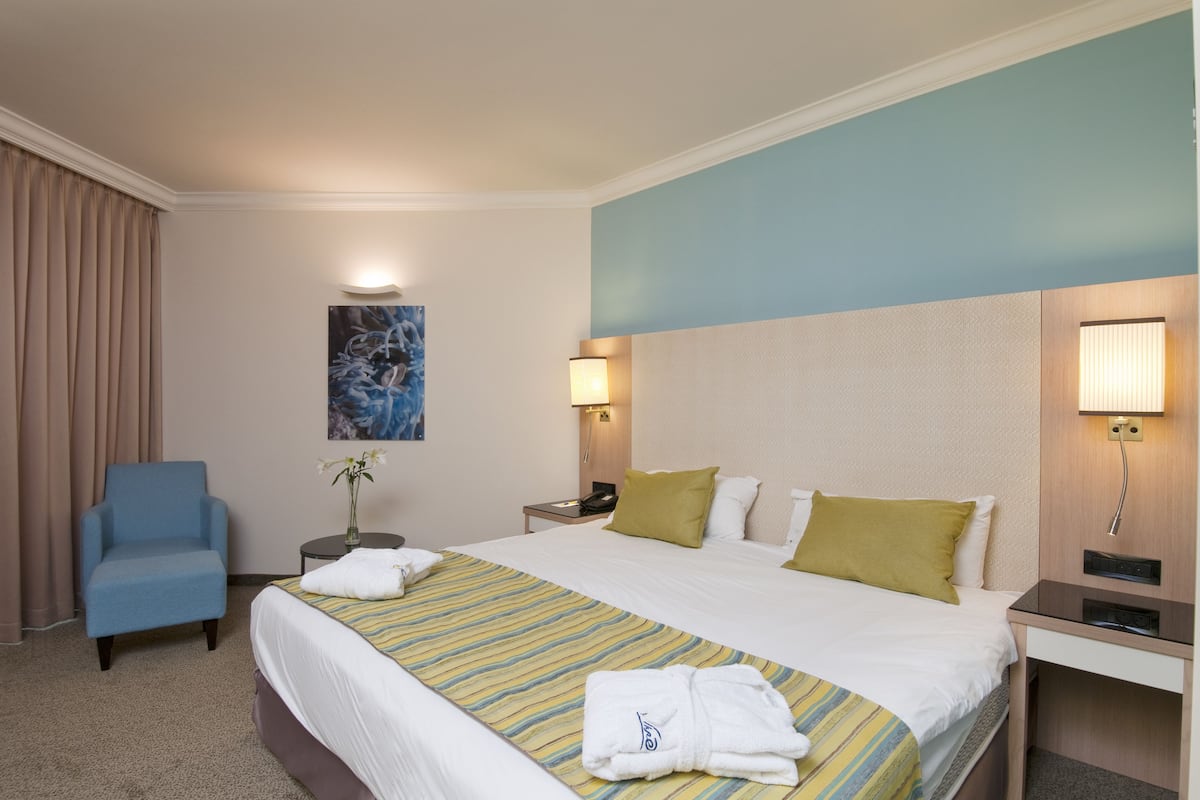 A hotel room with a large bed, a blue armchair, a bedside table with a lamp, and a painting on the wall.