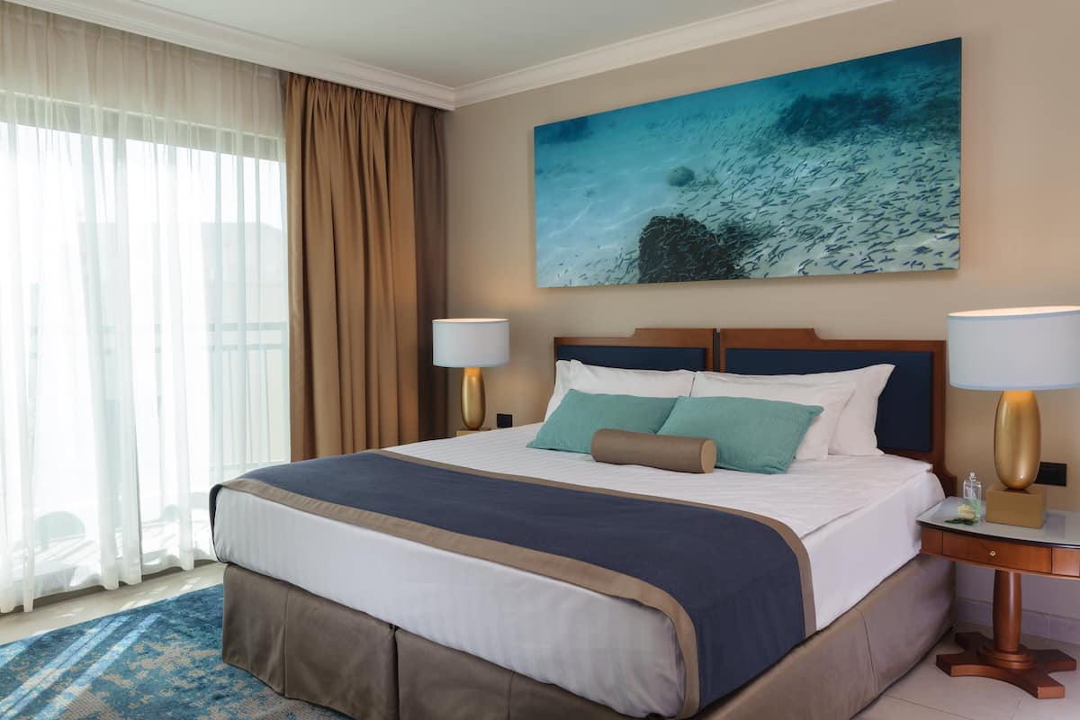 A hotel room with a large bed, a nightstand with a lamp, a painting of an underwater scene, and sheer curtains.