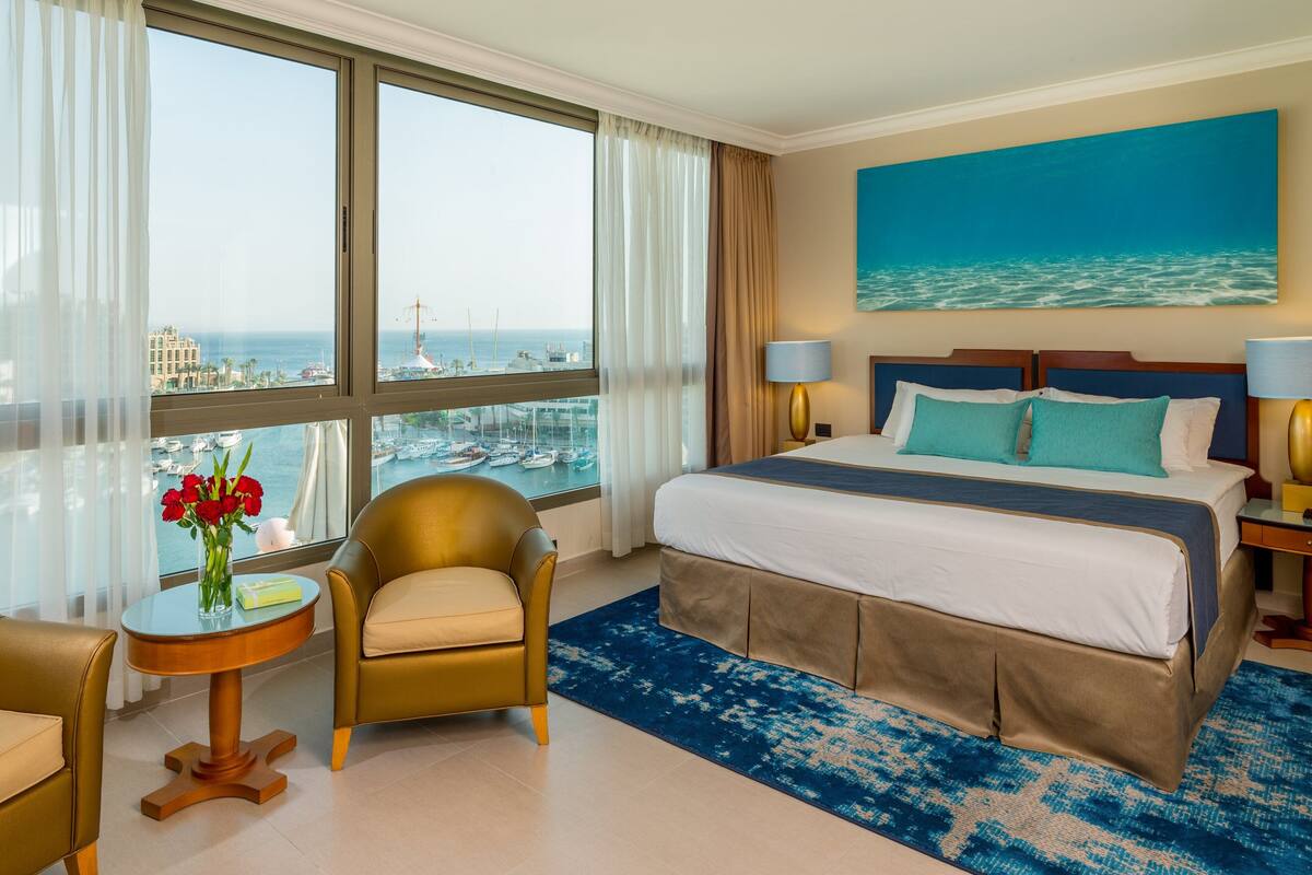 A hotel room with a large bed, a view of the sea, a round table with a vase of flowers, and a chair.
