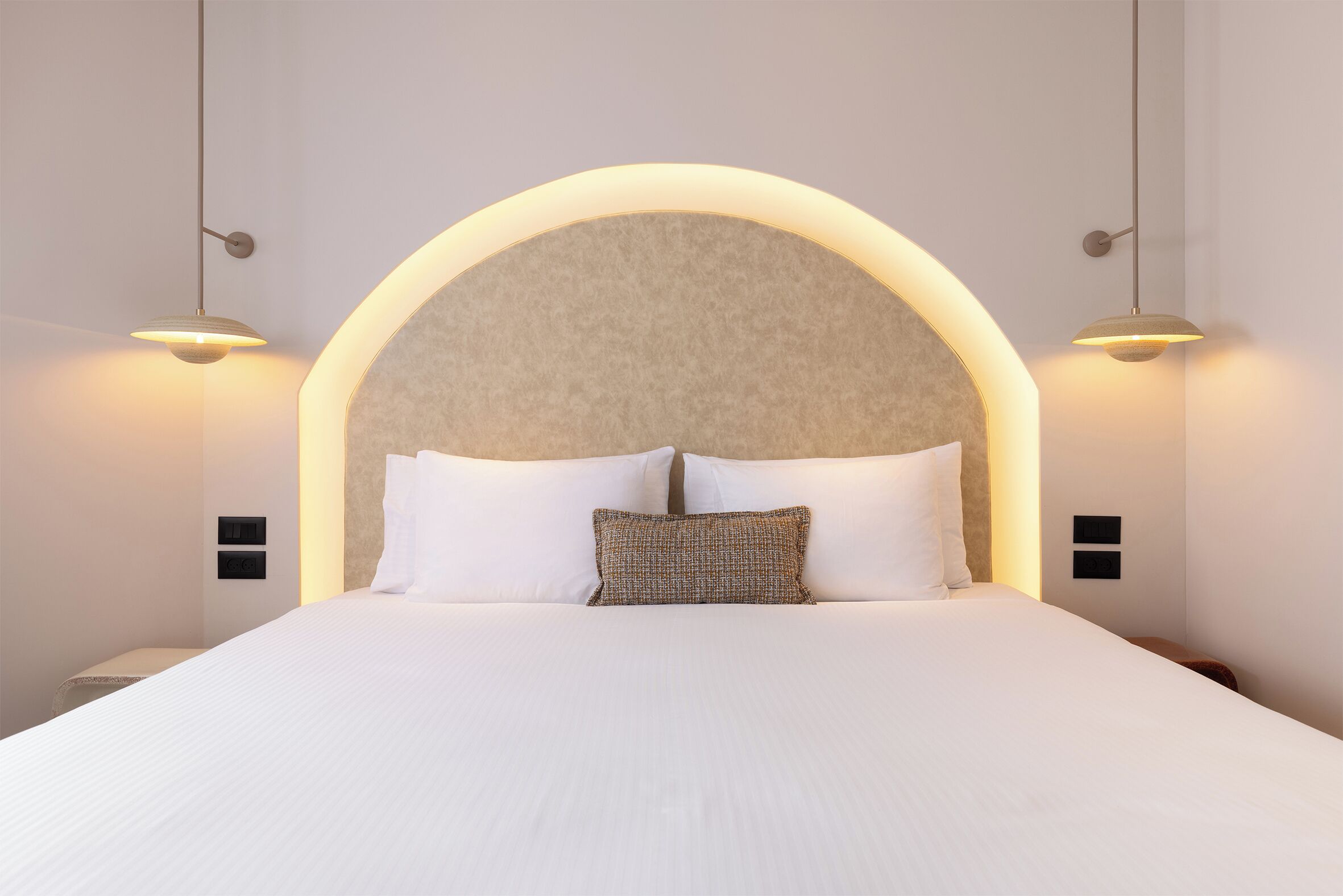 Una cama impecable con ropa de cama blanca y un almohadón texturizado, flanqueada por dos lámparas de pared.
