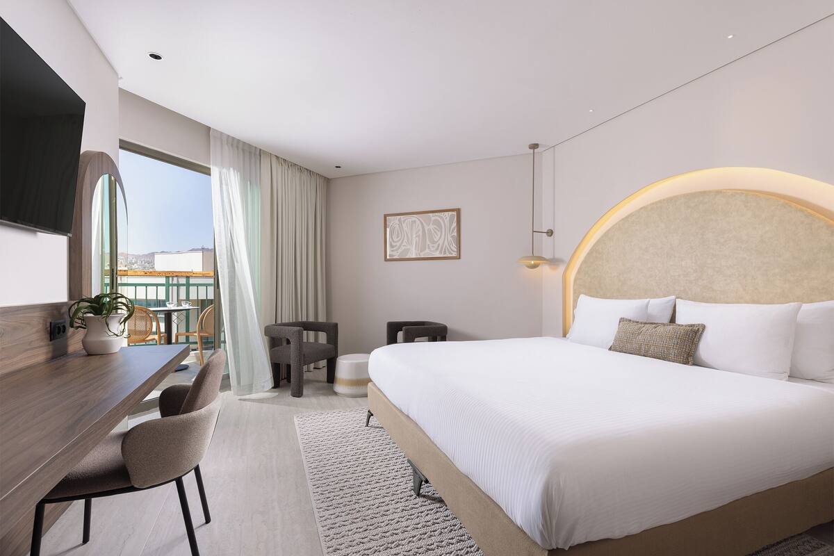 A modern hotel room with a large bed, a desk with a chair, a small table, and a potted plant.