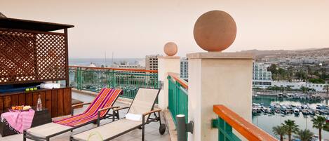 A balcony with lounge chairs, a wooden table with fruits, and a view of a marina and cityscape.