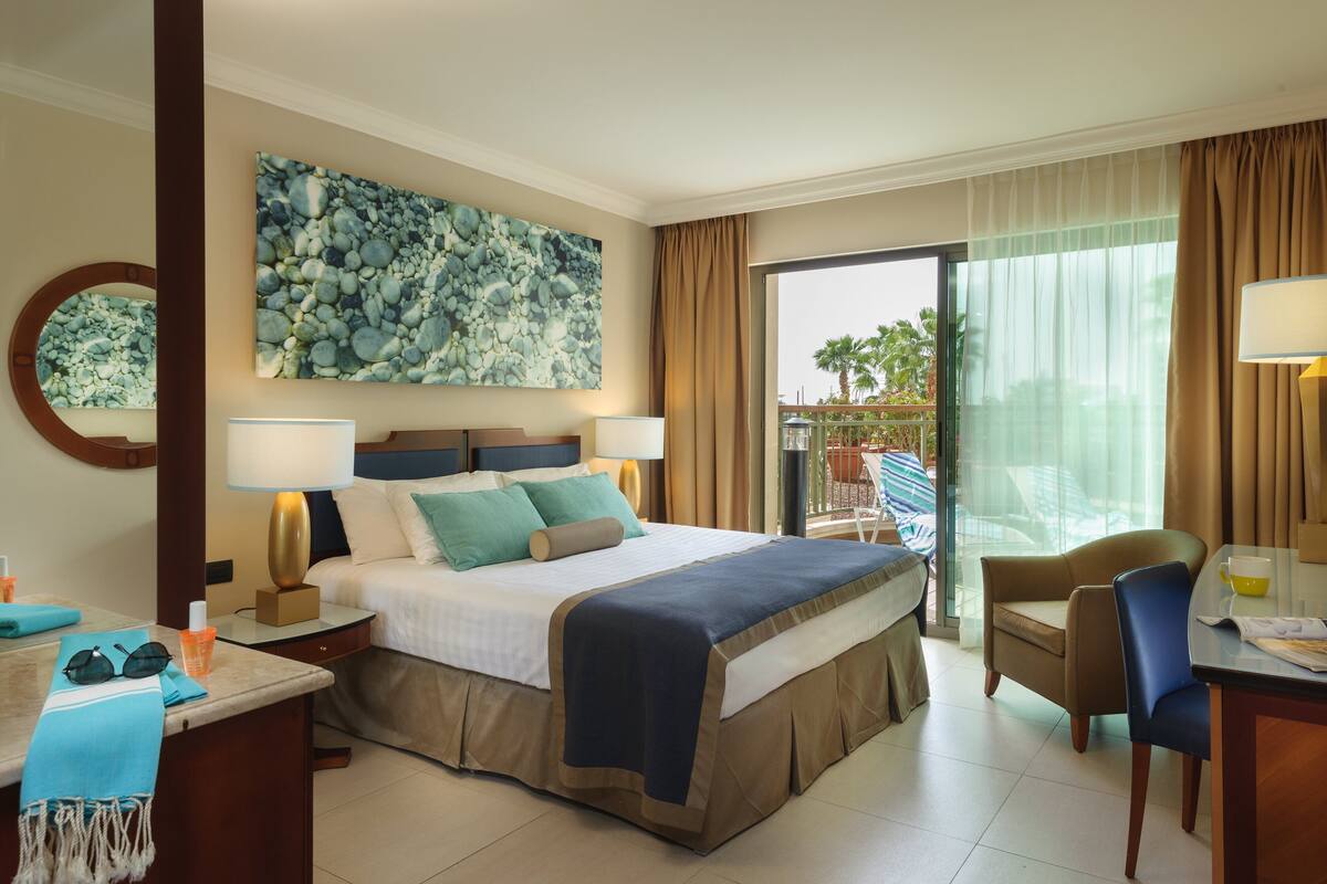 A hotel room with a bed, bedside lamps, a desk with a chair, a large window with curtains, and a view of the outdoors.