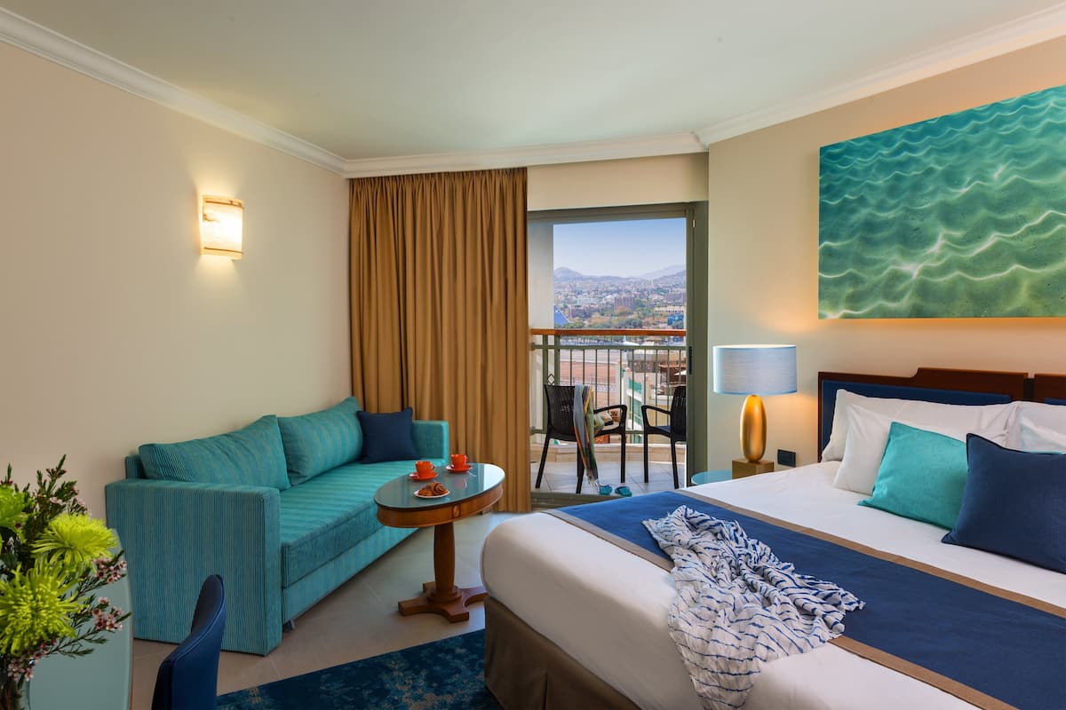 A hotel room with a bed, a sofa, a small table with a cup and saucer, a chair, and a view of the city through a large window.