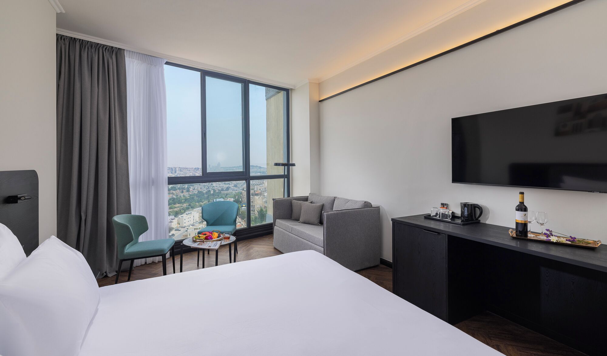 A hotel room with a large bed, a desk with a TV, a small table with a newspaper, a chair, and a view of the city through large windows.