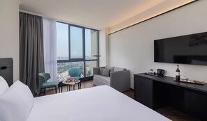 A hotel room with a large bed, a desk with a TV, a small table with a newspaper, a chair, and a view of the city through large windows.