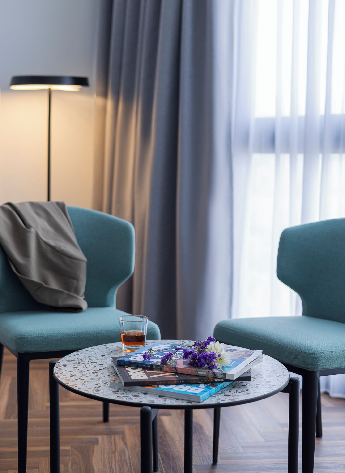 Een moderne kamer met twee turquoise fauteuils, een ronde salontafel met tijdschriften en bloemen, een kop thee en een lamp.