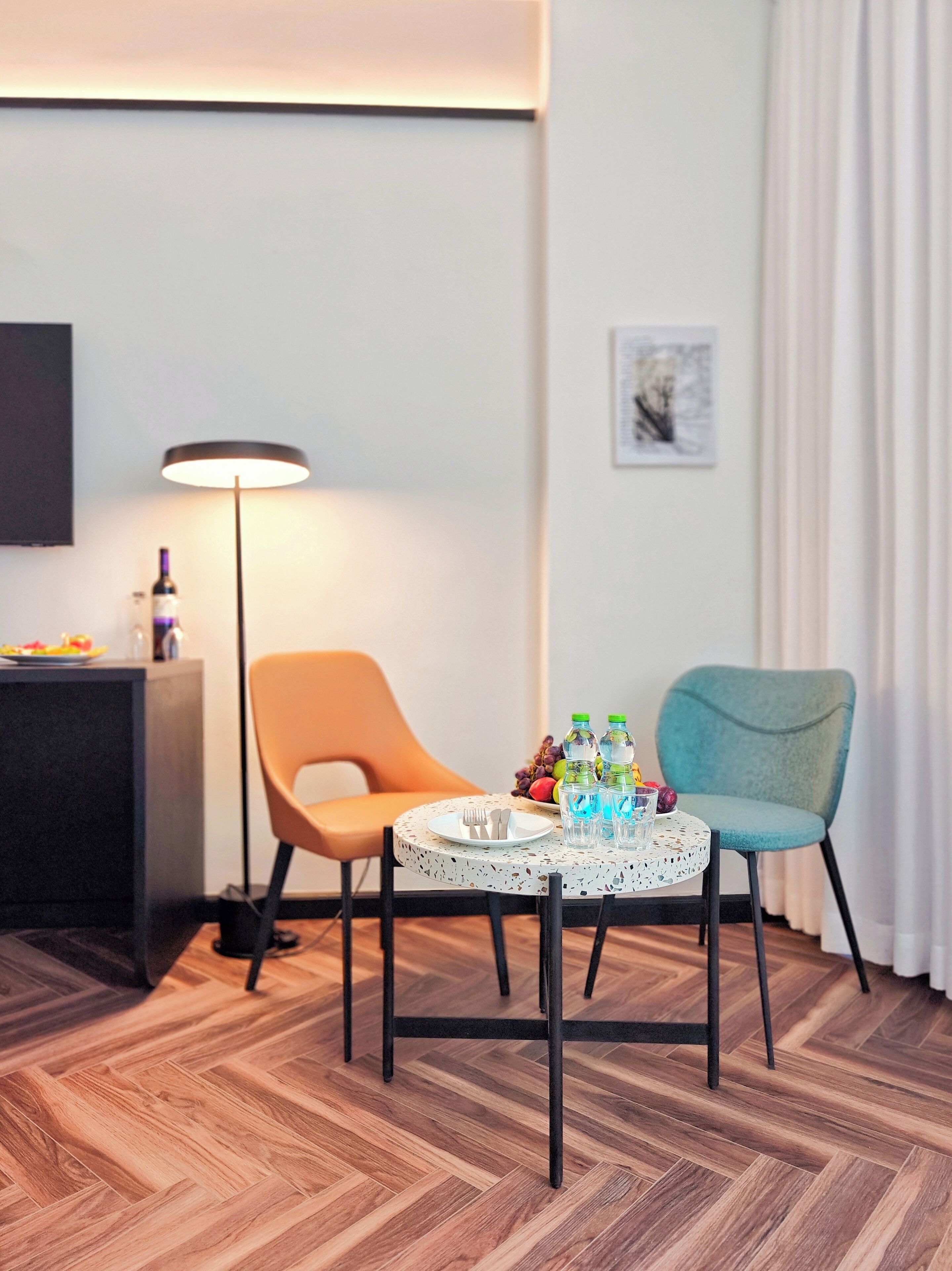 Ein Hotelzimmer mit rundem Tisch, zwei Stühlen, einer Lampe, einem kleinen Schrank und einem an der Wand befestigten Fernseher.