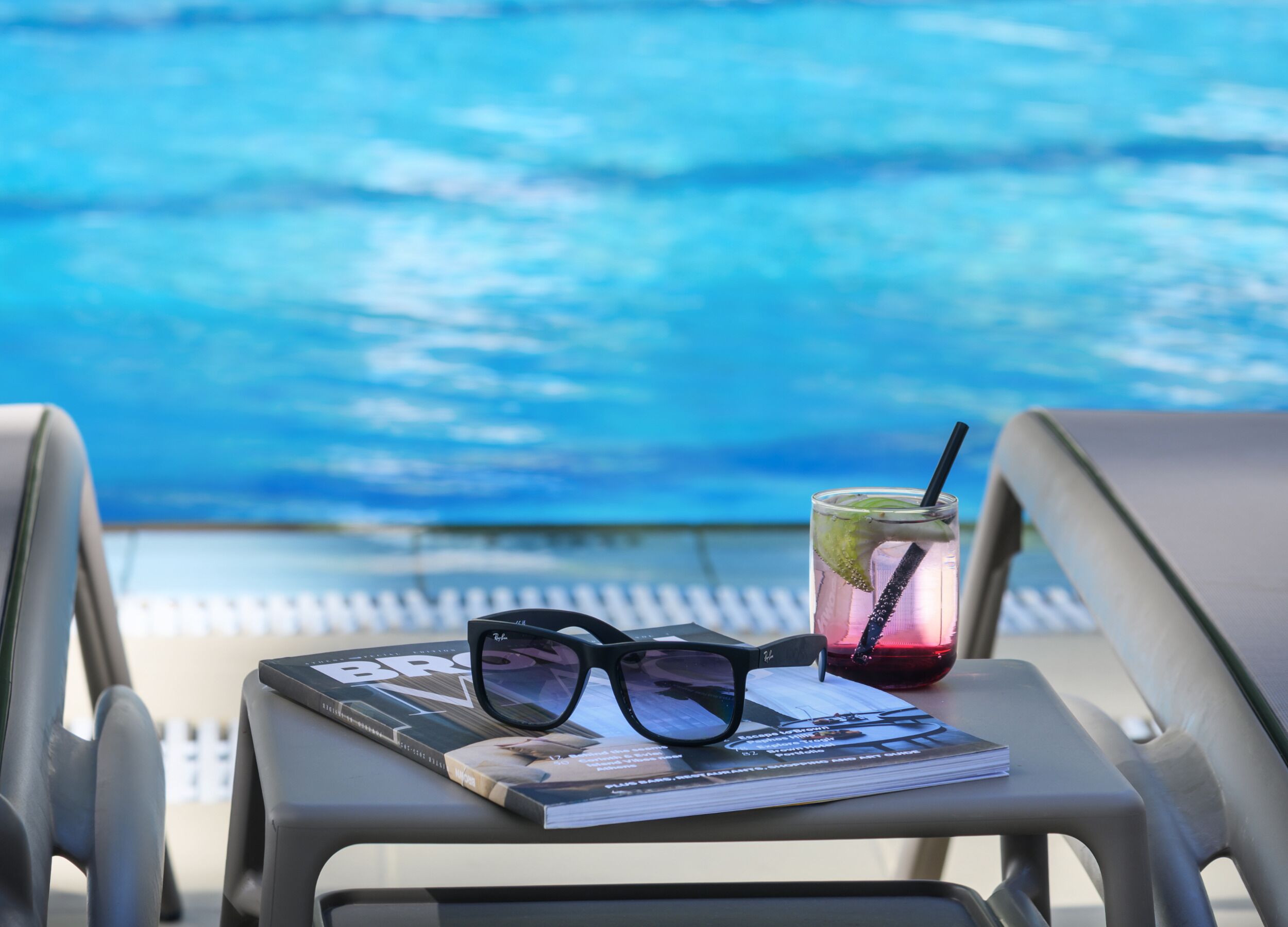 A pair of sunglasses, a drink with a straw, and a magazine on a chair by a pool.