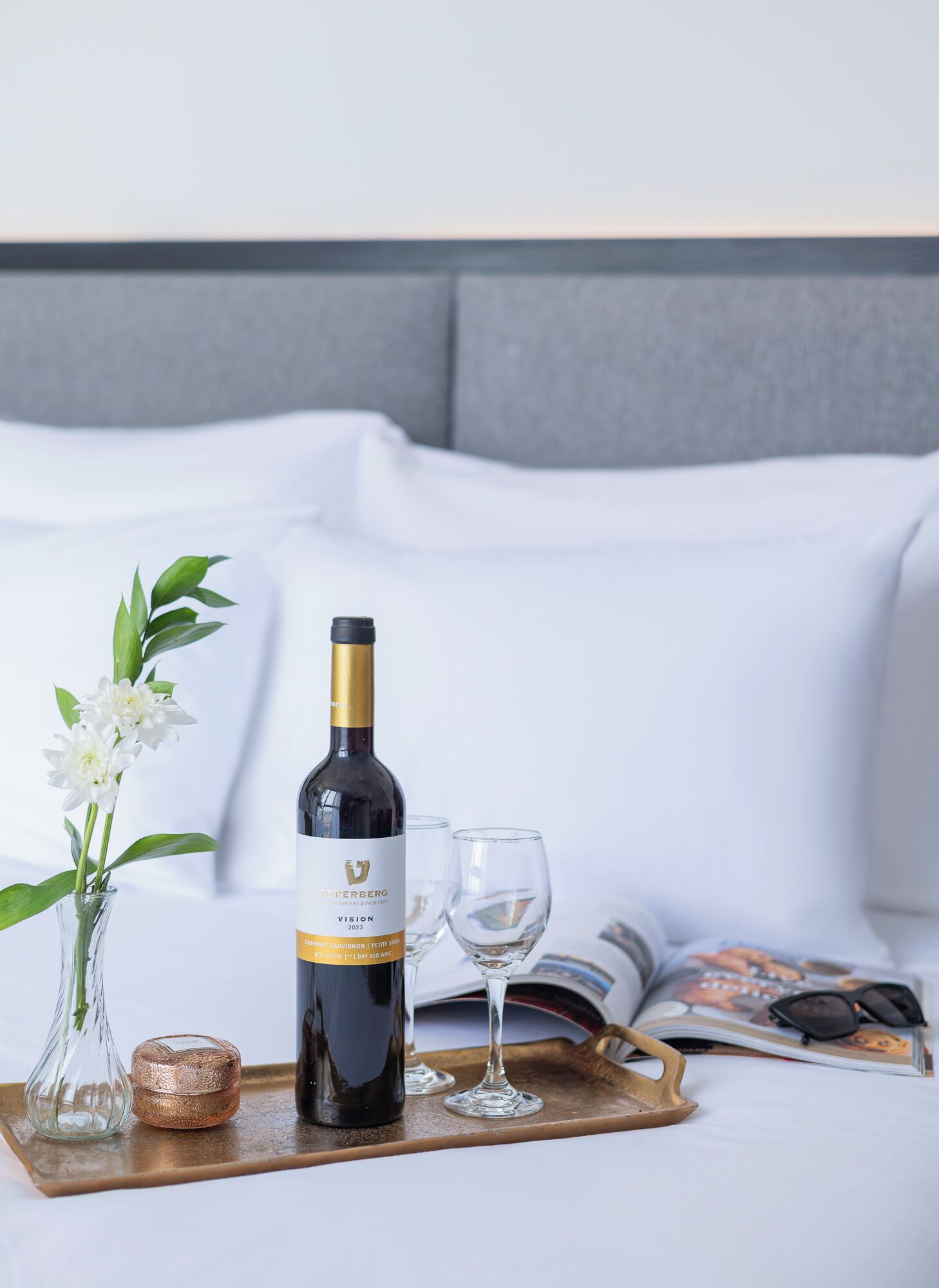 Una bottiglia di vino, due bicchieri, un vassoio, un libro e un vaso con fiori su un letto.