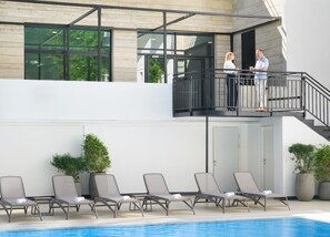 Un bâtiment moderne doté d’une piscine, de chaises longues et d’un balcon où peuvent s’asseoir deux personnes.