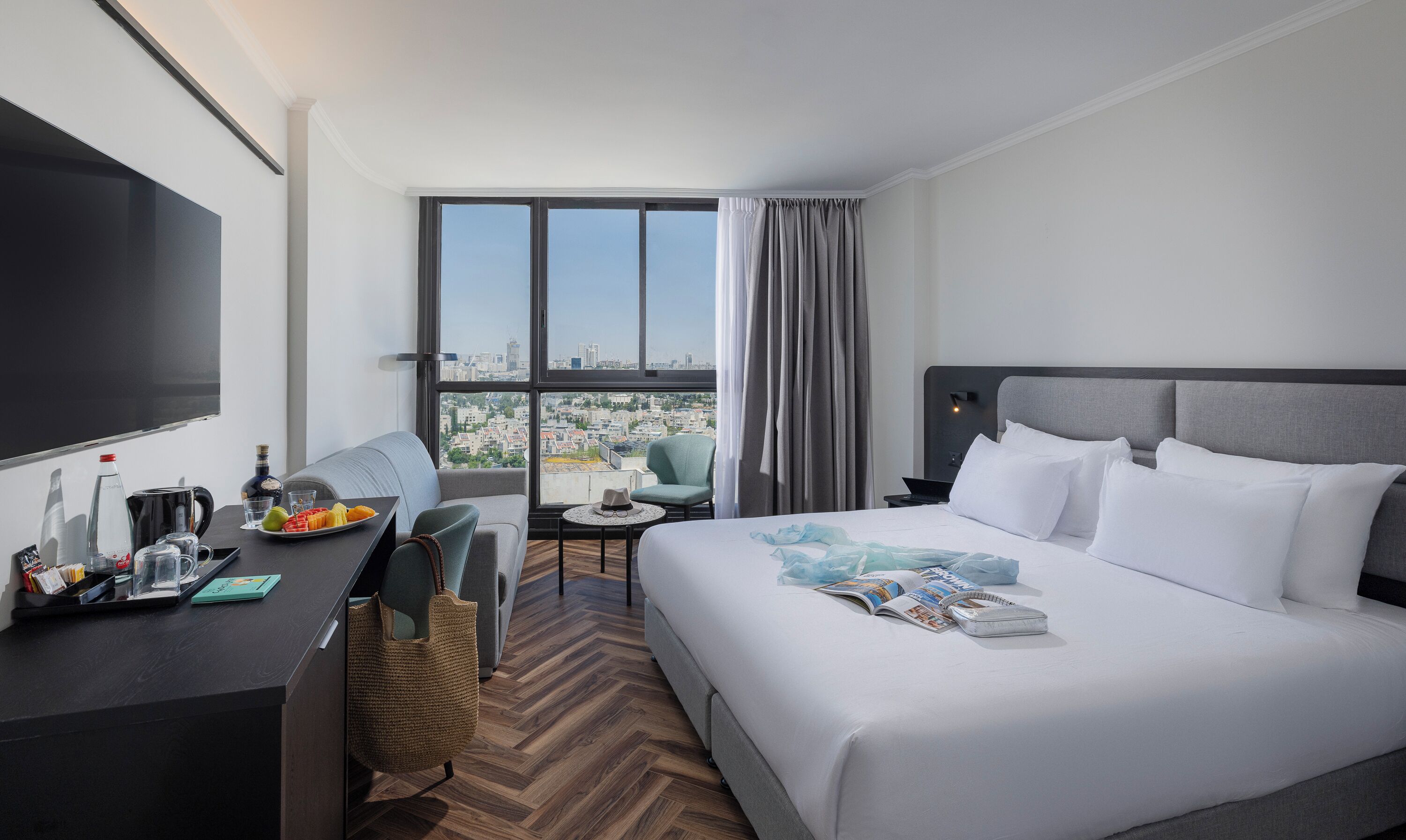 A hotel room with a large bed, a desk with a flat-screen TV, a chair, a small table, and a view of the city through large windows.